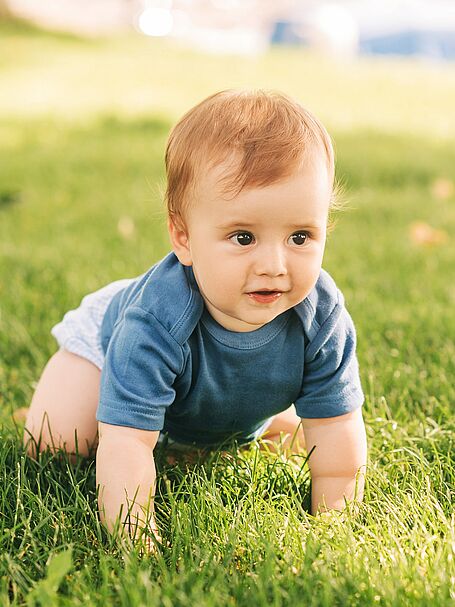 Baby crawling on the grass in a park, wearing a blue shirt and looking curious.